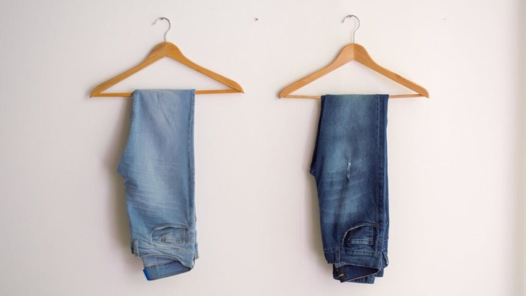 Twee jeansbroeken in licht- en donkerblauw hangen gevouwen aan houten kledinghangers, symbool voor slim fit vs tapered fit